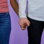 Two people, one wearing a red and white striped shirt and blue jeans, the other a white long-sleeved shirt and black pants, hold hands against a lavender background.