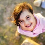 A young girl with windblown hair smiles up at the camera, wearing a pink hoodie.