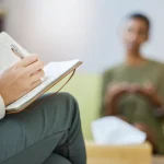 A therapist's hand writes in a notebook while a client is in the background, out of focus.