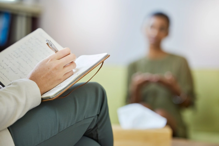 A therapist's hand writes in a notebook while a client is in the background, out of focus.